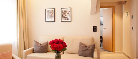 A modern living room with a beige sofa, a glass coffee table with magazines, and a vase of red roses. There are two framed pictures on the wall and a wooden door in the background.