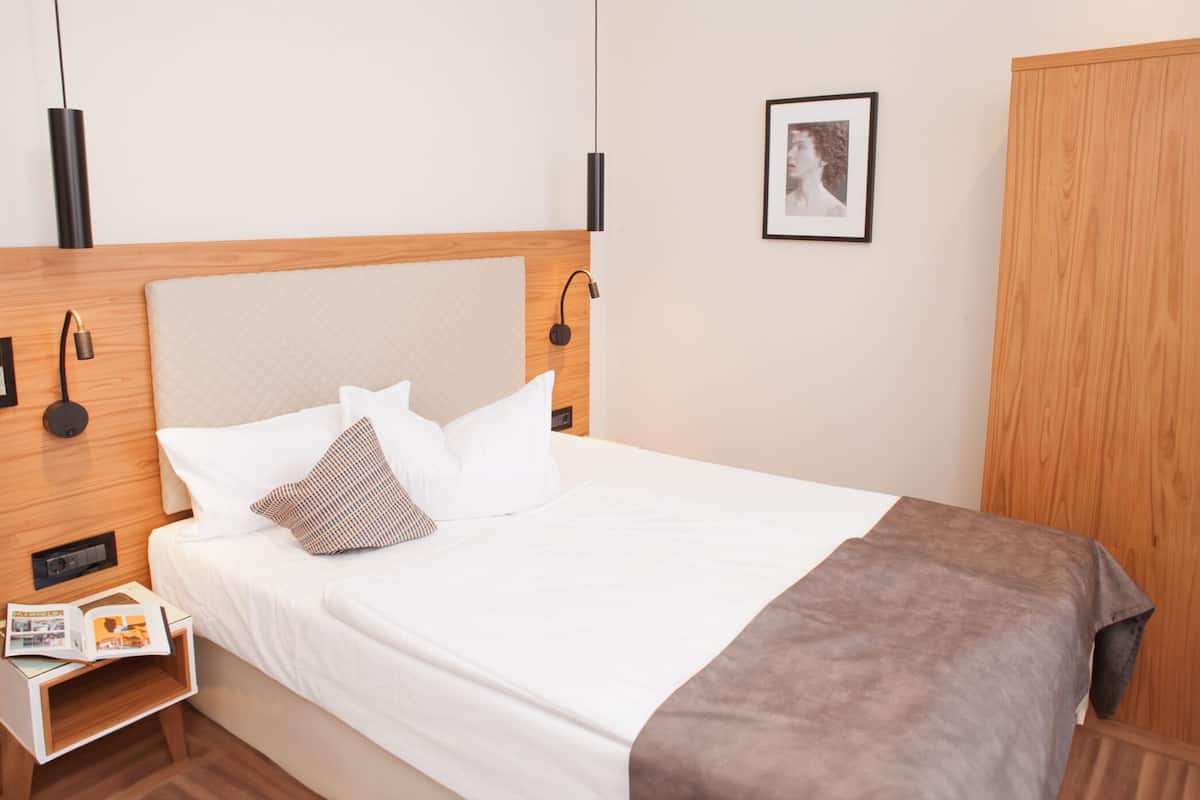 Una cama bien hecha con almohadas blancas y estampadas, un cabecero de madera, una mesita de noche con una revista y un cuadro en la pared.