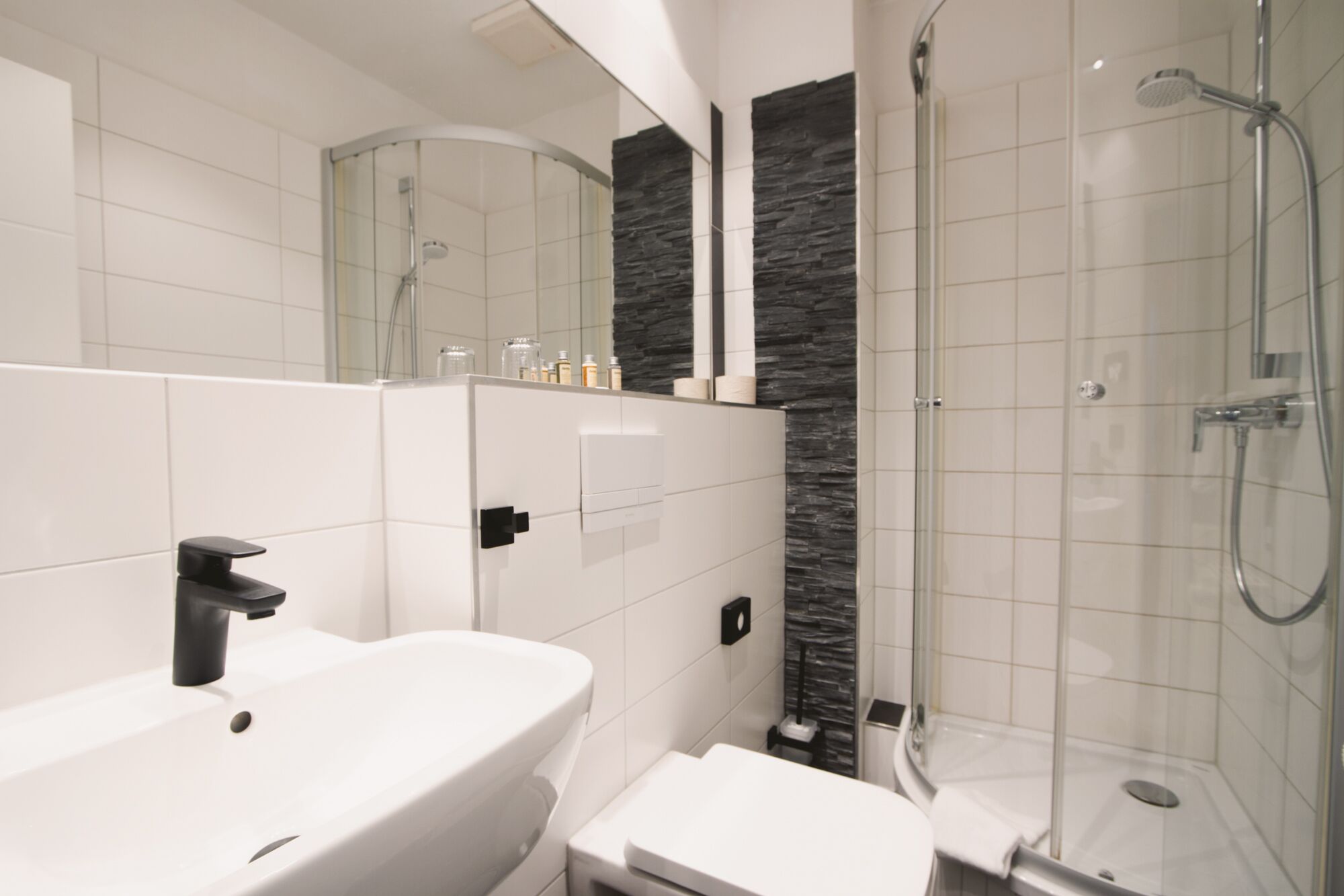 A modern bathroom with a white sink, a mirror, a glass shower enclosure, and a stone accent wall.