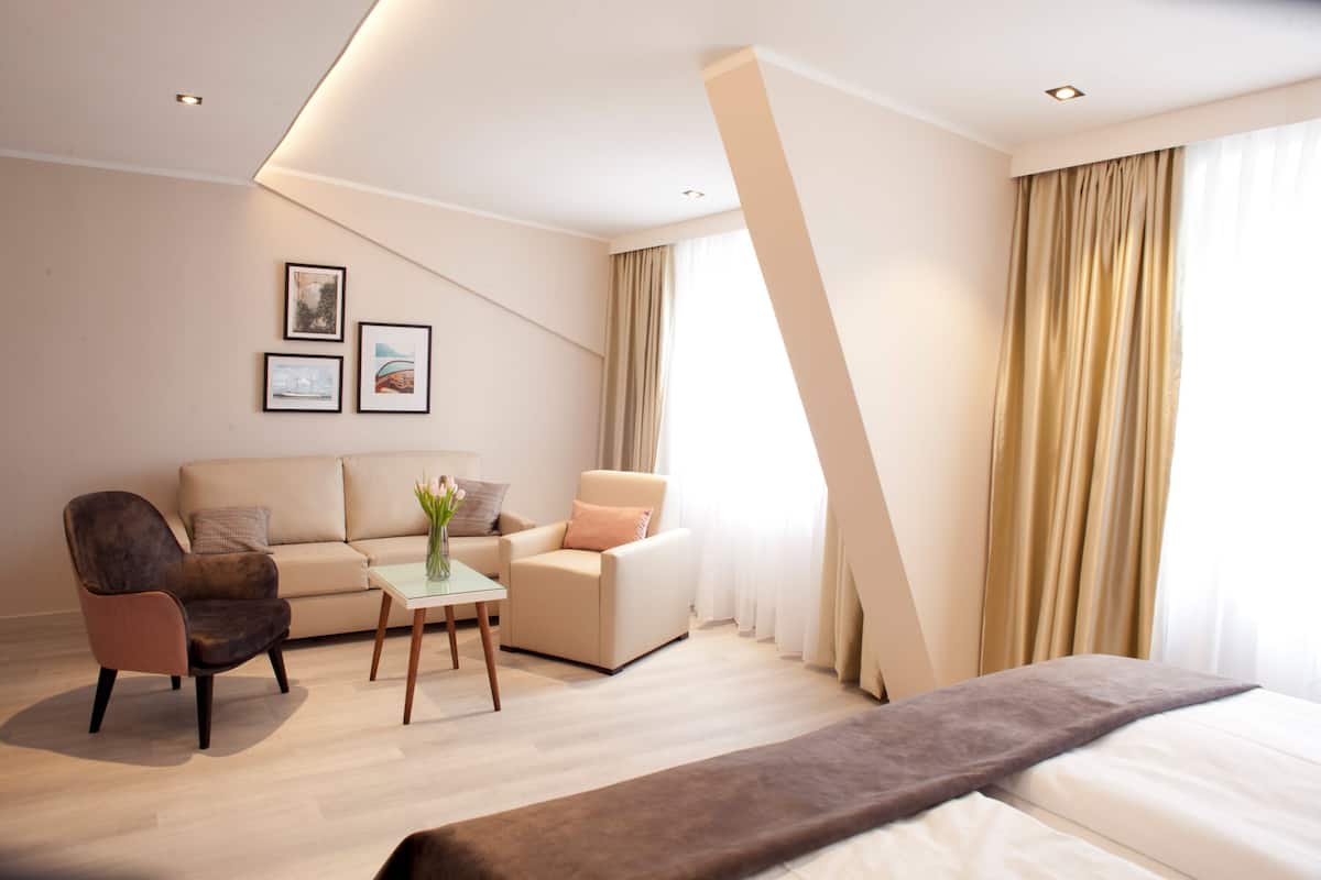 Una habitación de hotel moderna con una cama, un sofá, una silla, una mesita pequeña y una mesa de centro. Hay dos cuadros enmarcados en la pared.