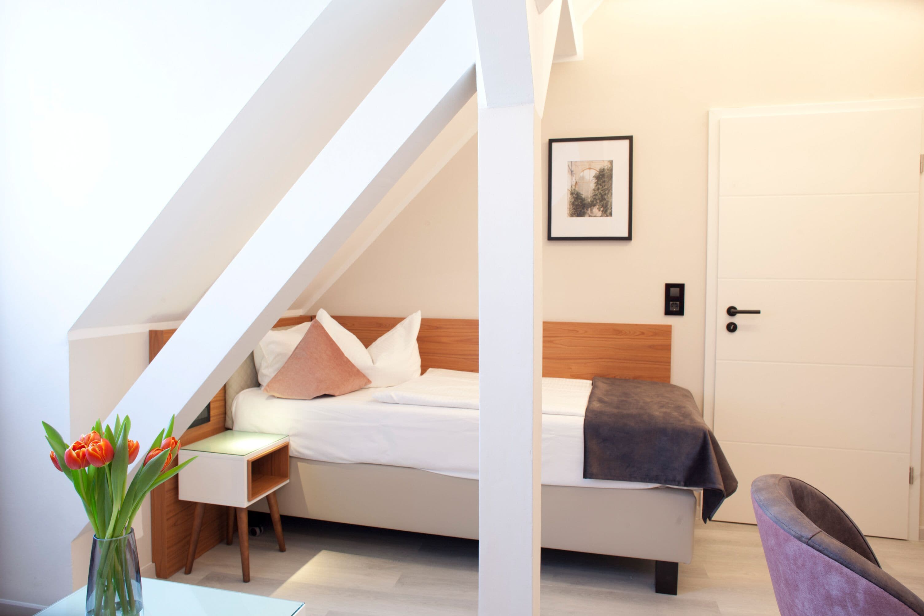 A bedroom with a bed, a nightstand, a chair, a door, and a framed picture on the wall.