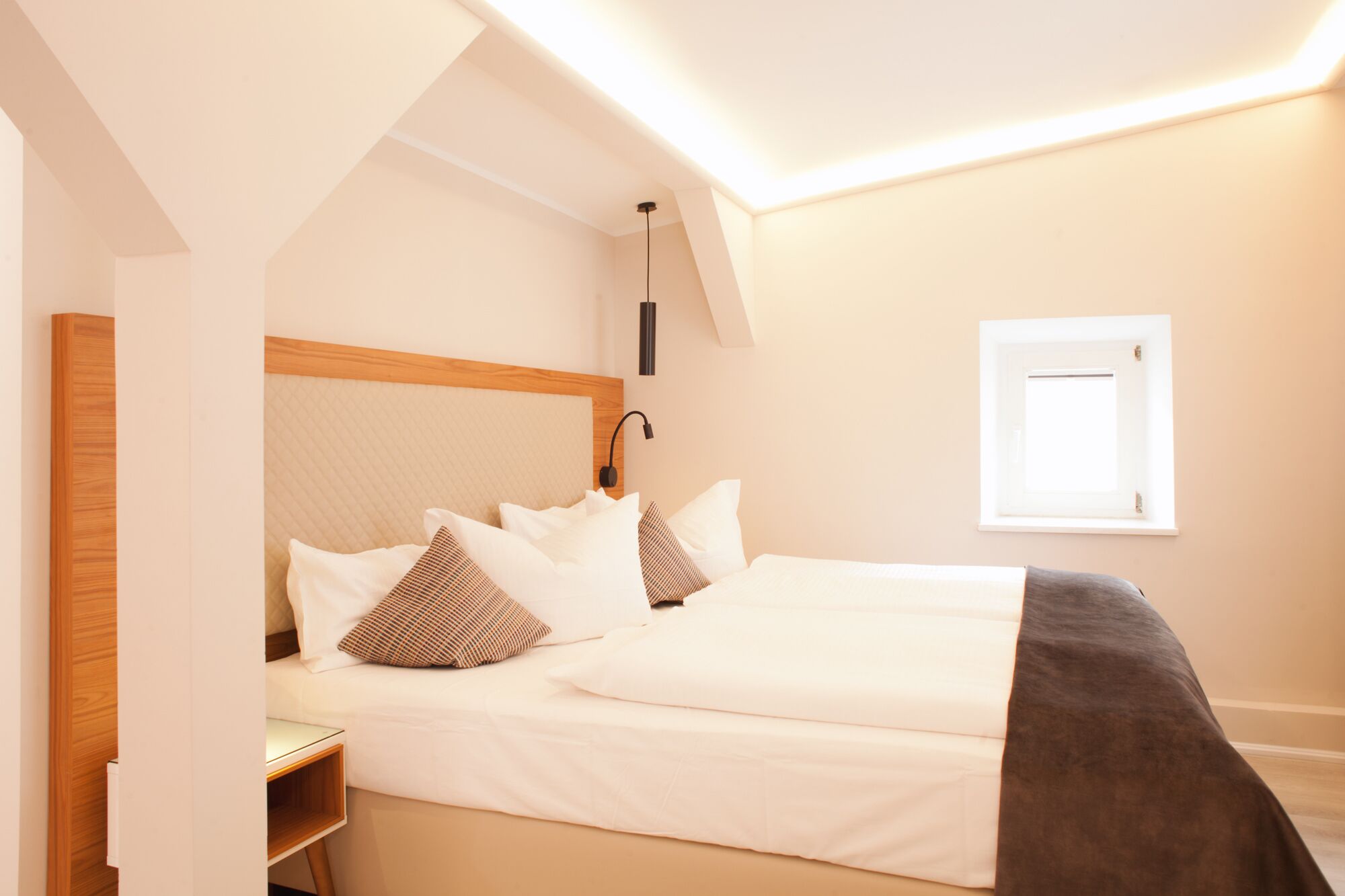 Quarto com cama, almofadas, mesa de cabeceira, abajur e janela.
