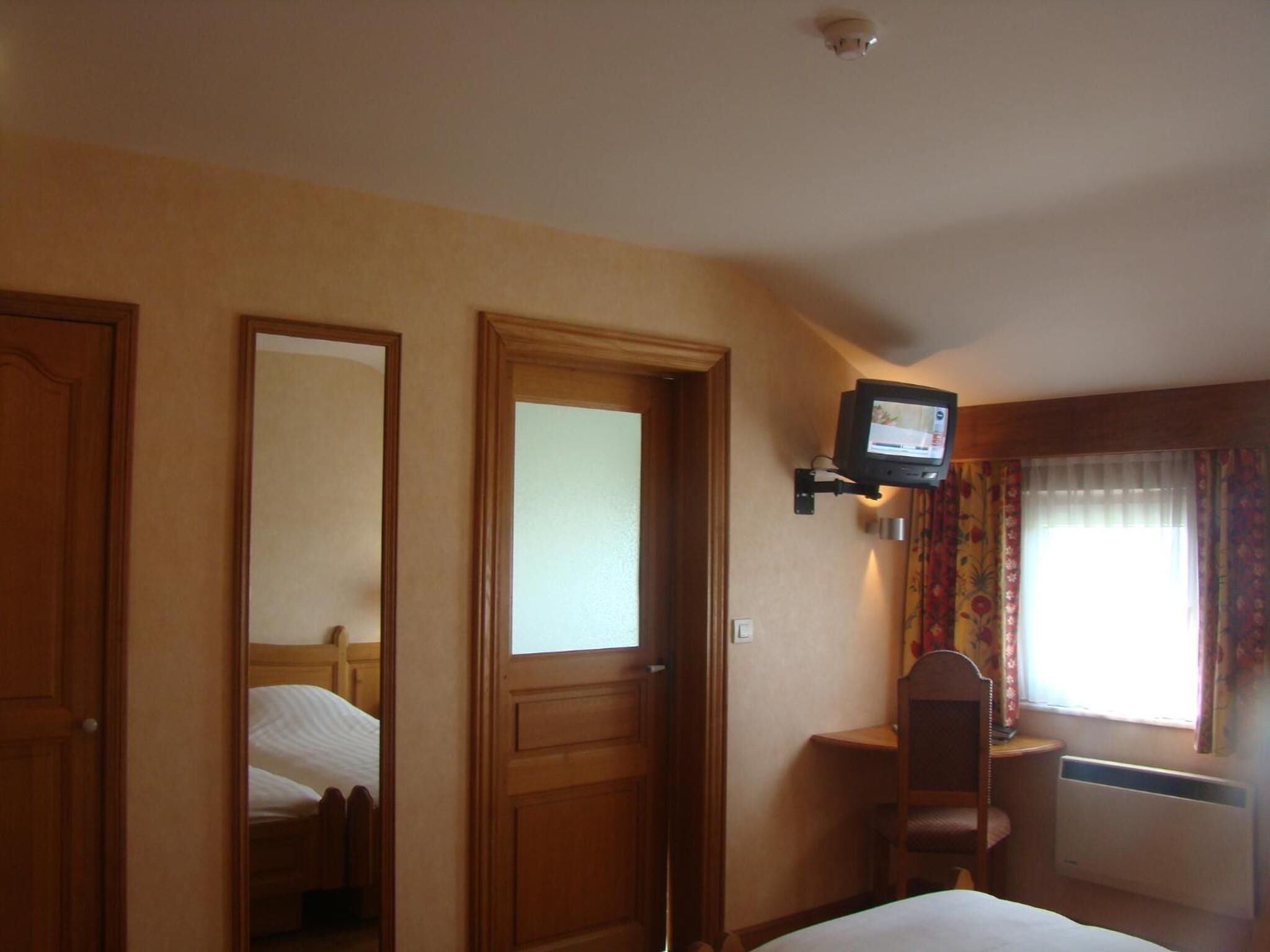 Habitación de hotel con cama, una silla, televisor montado en la pared y ventana con cortinas.