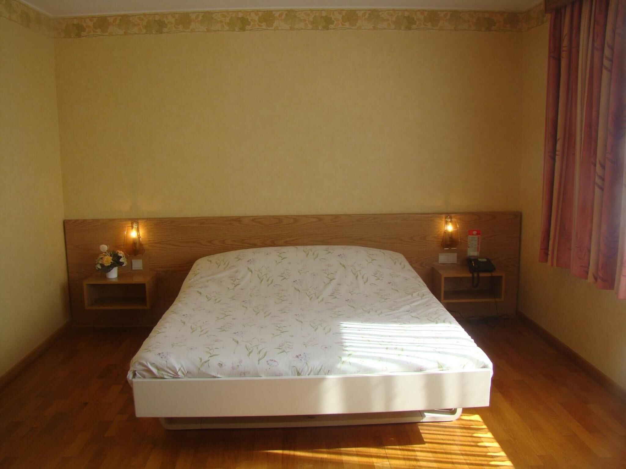 A neatly made bed in a room with wooden floors and a plain yellow wall. There are two bedside tables with lamps and a small decorative item on each.