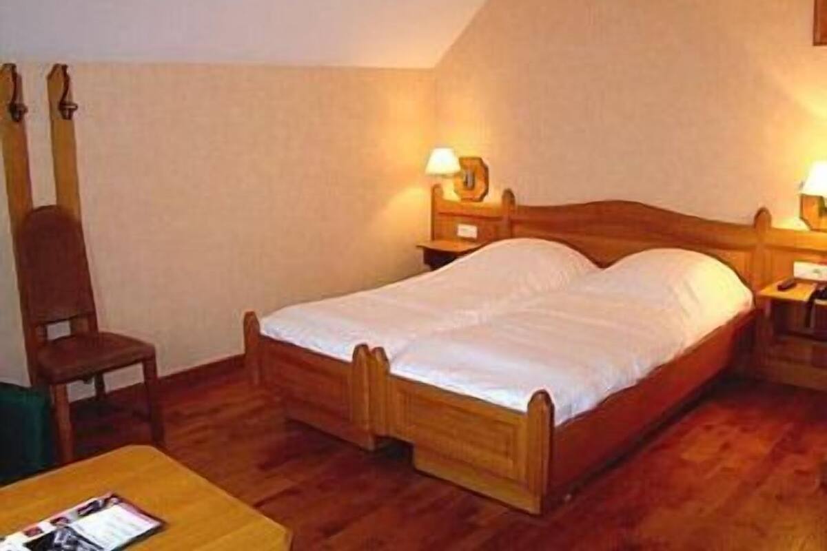 Un lit simple avec une tête de lit et un pied de lit en bois, deux tables de chevet avec des lampes, une chaise en bois et une petite table avec un magazine.