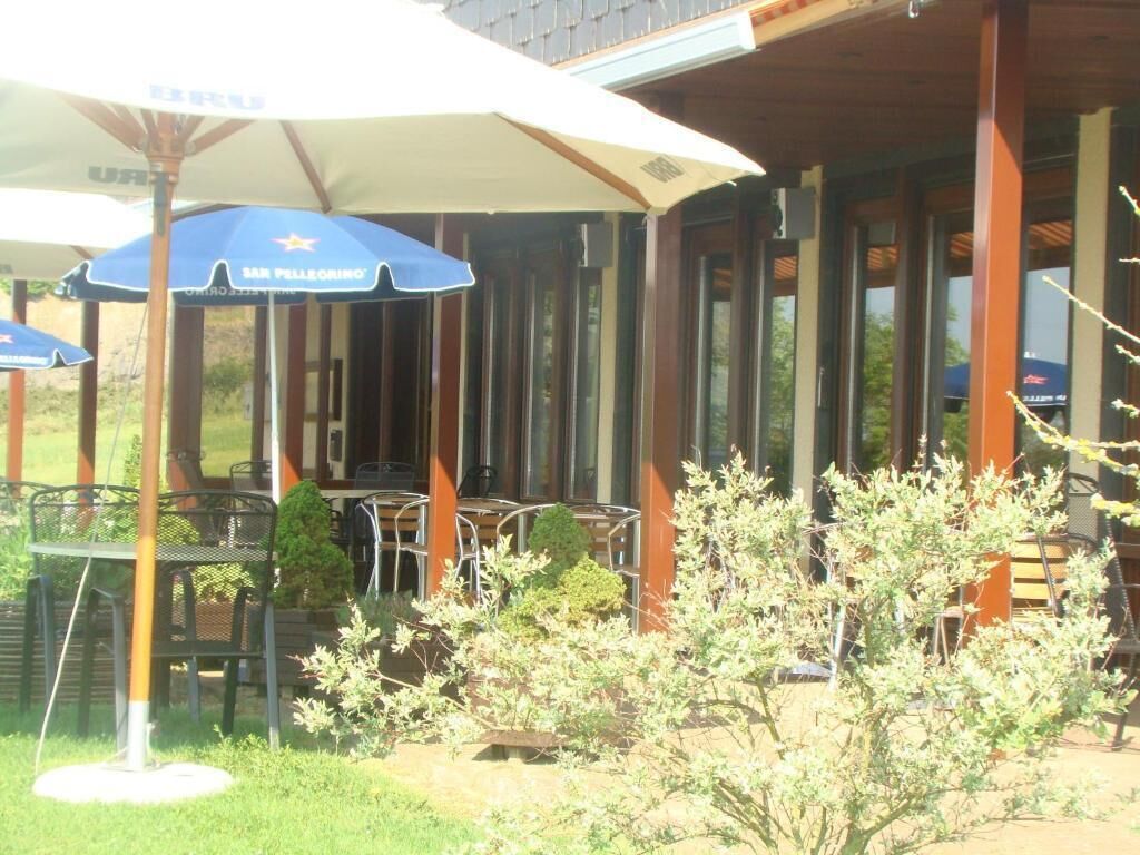 Ein Terrassenbereich im Freien mit Tischen und Stühlen unter Sonnenschirmen, umgeben von Holzsäulen und Glastüren.