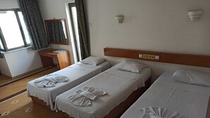 A hotel room with two single beds, each with a towel and pillow. There is a small desk with a chair, a window with curtains, and a wall-mounted light fixture. - Dias (Kusadasi)