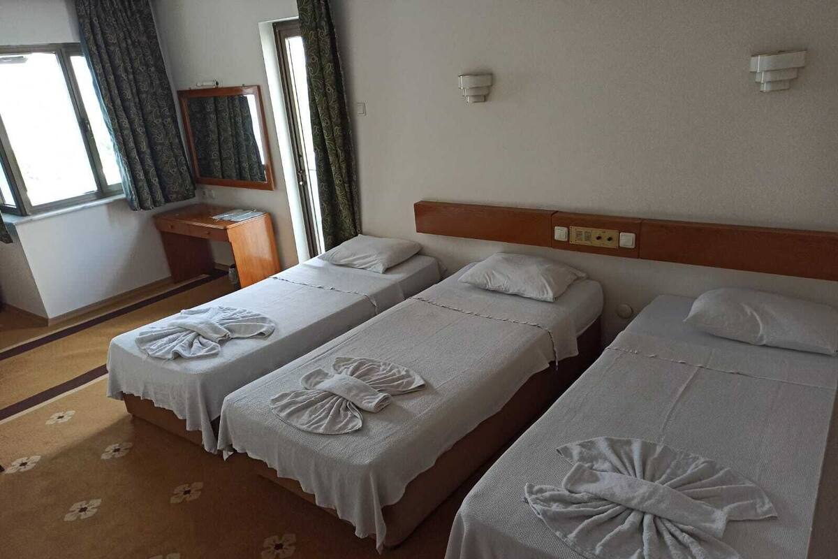 A hotel room with two single beds, each with a towel and pillow. There is a small desk with a chair, a window with curtains, and a wall-mounted light fixture.