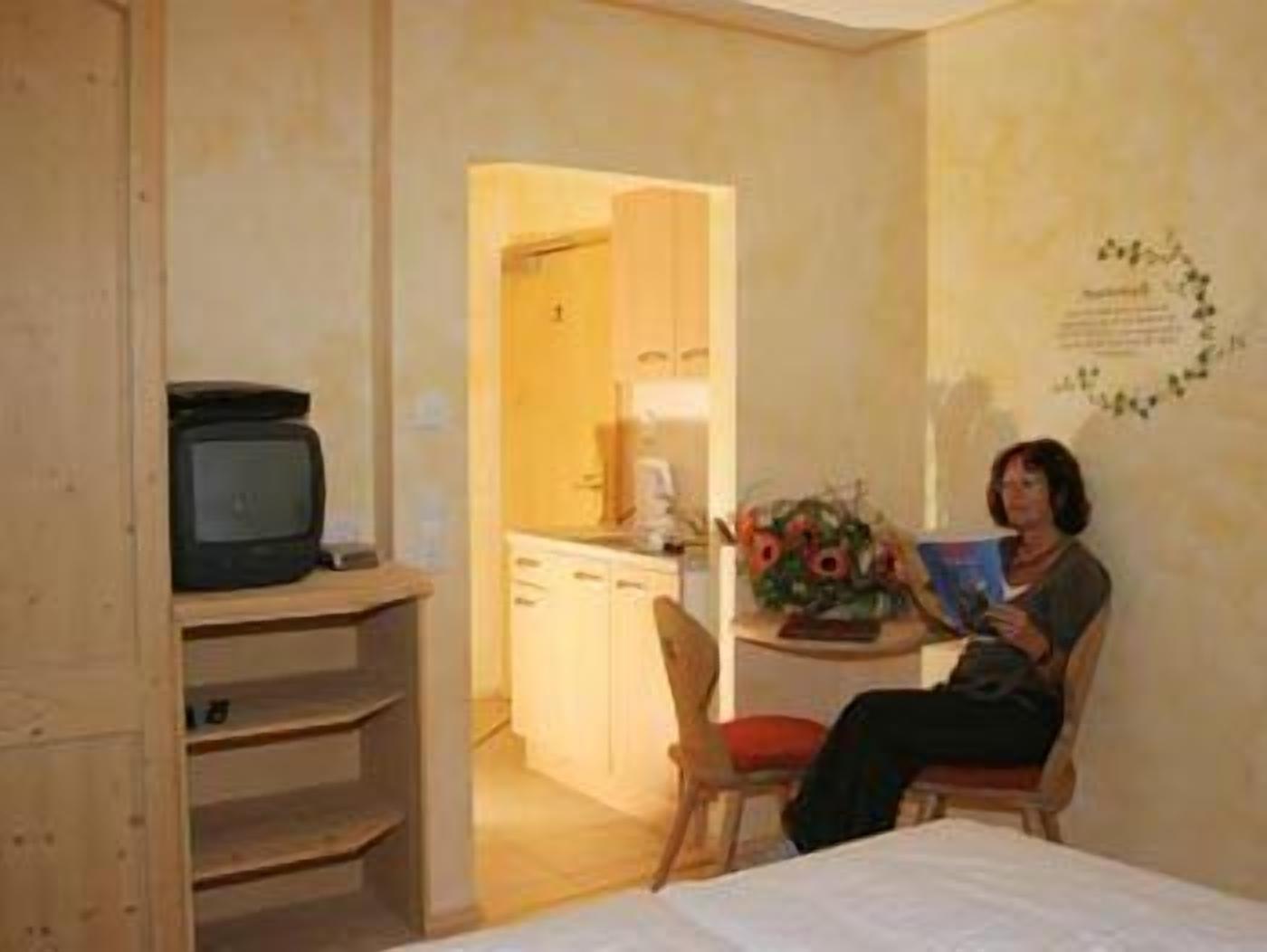Ein kleines Zimmer mit einem Bett, einem Tisch mit Stuhl, einem Fernseher auf einem Regal und einer sichtbaren Küchenzeile durch einen offenen Durchgang.