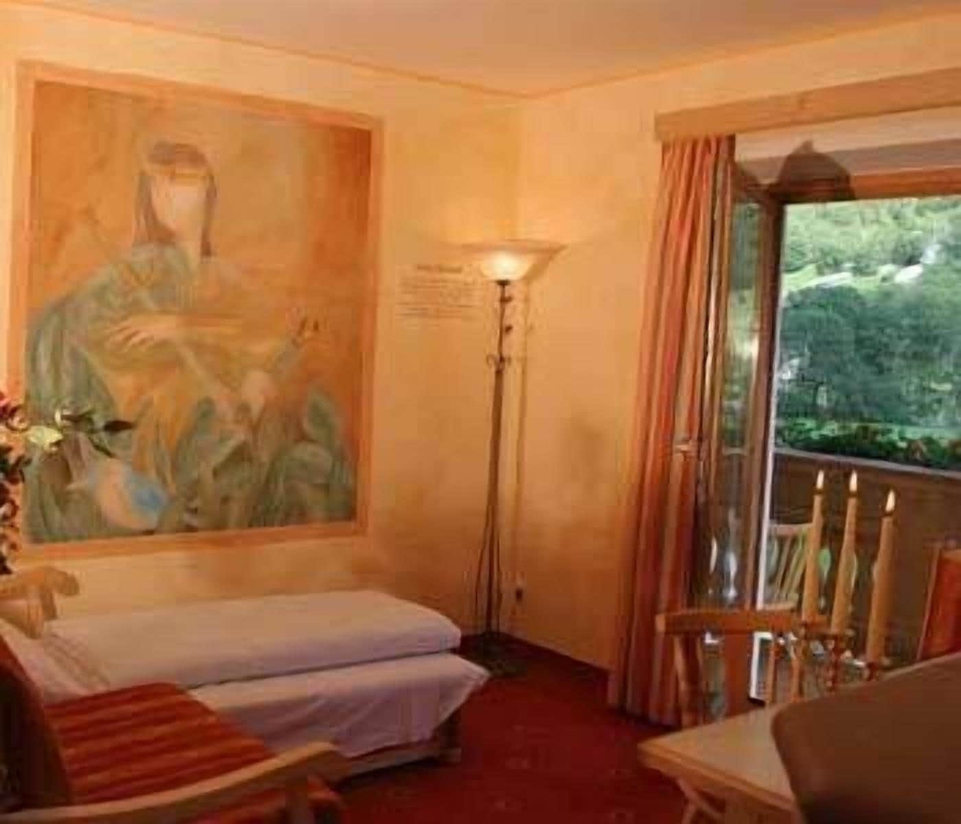 A hotel room with a bed, a painting on the wall, a floor lamp, and a view of greenery through the window.