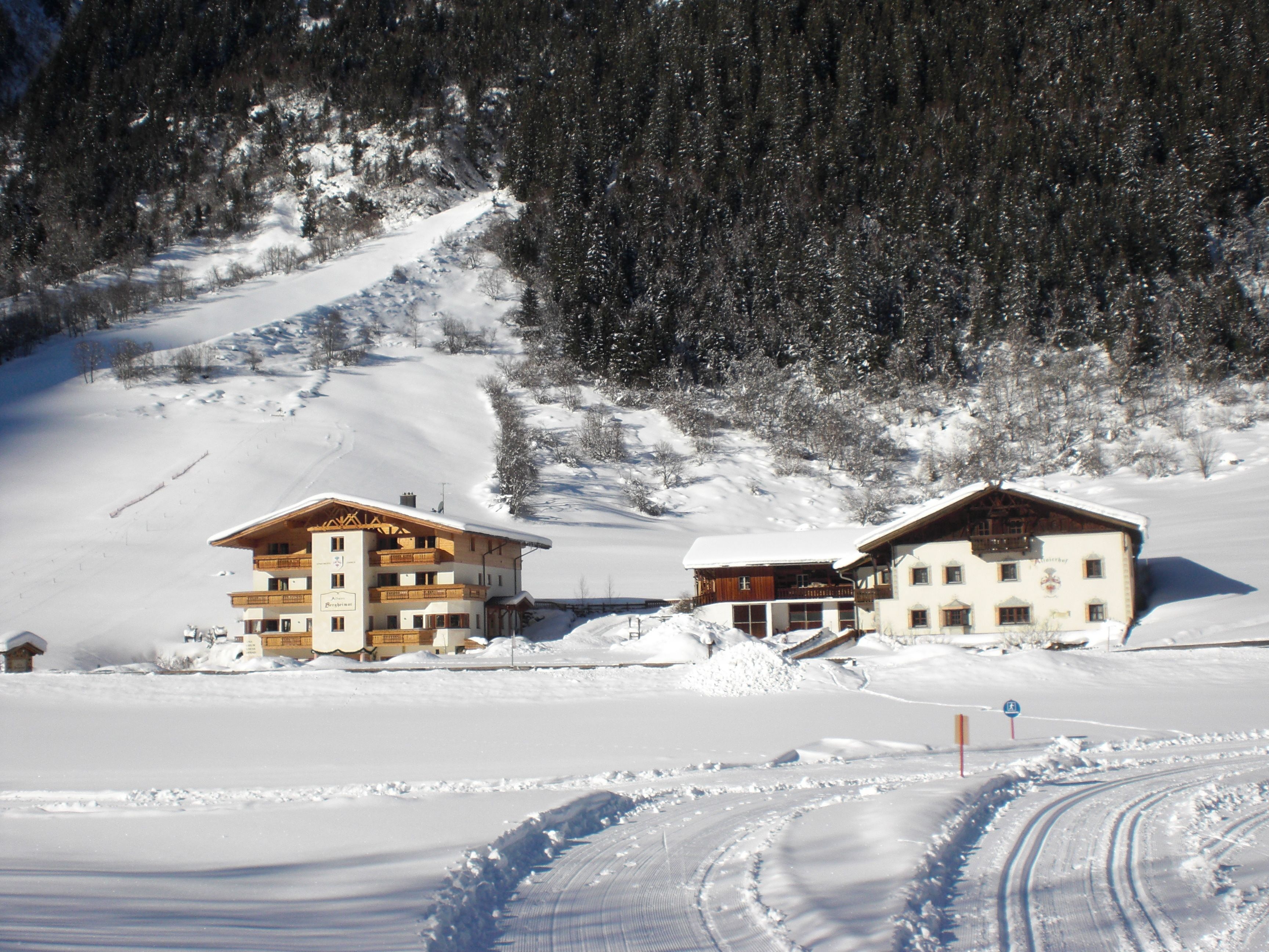 A snow-covered mountain area with two wooden buildings, likely a hotel, with visible balconies and a ski slope in the background.