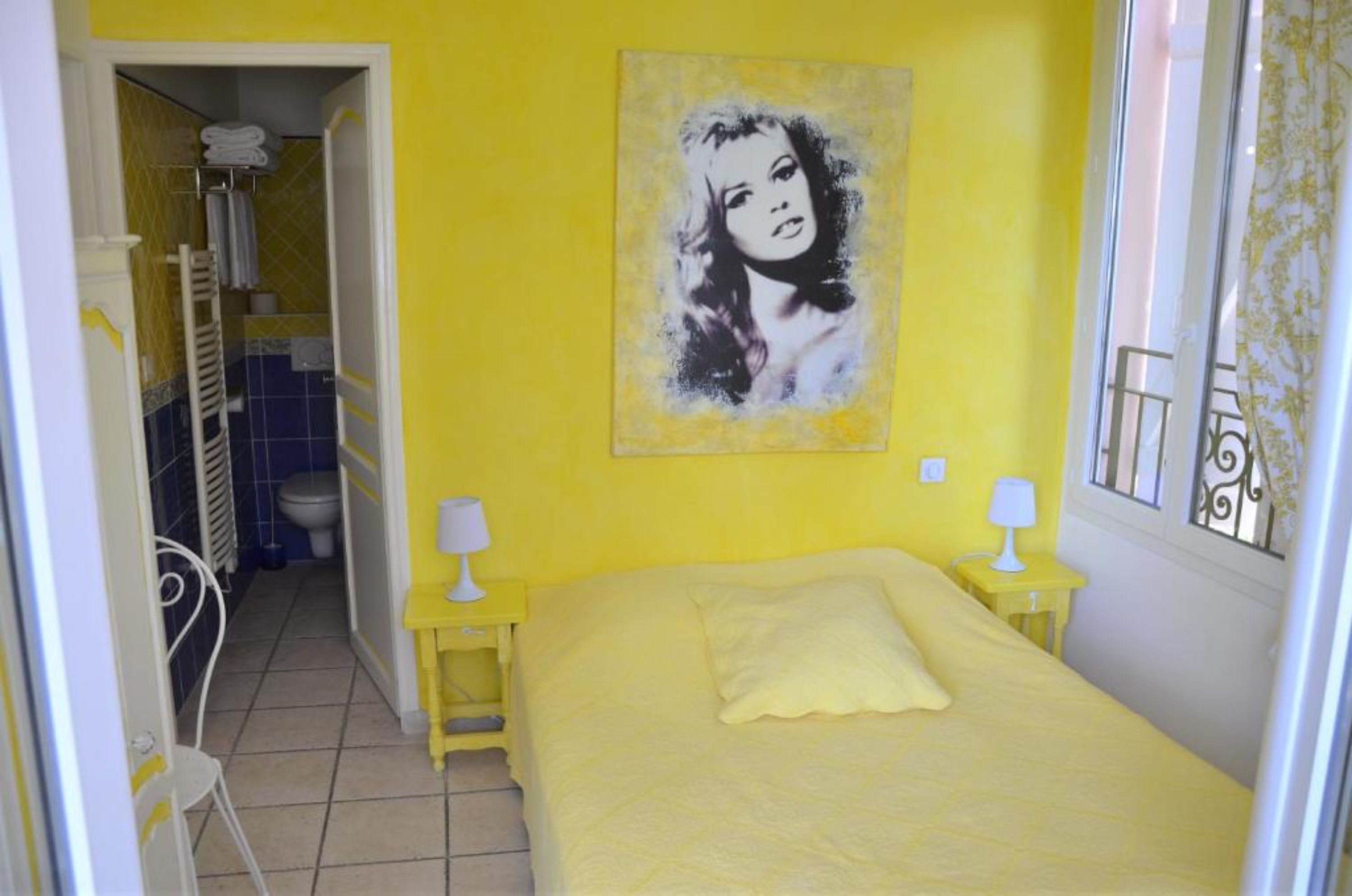 Habitación de color amarillo con una cama, dos mesitas de noche con lámparas, un baño visible a través de una puerta abierta y un cuadro en la pared.