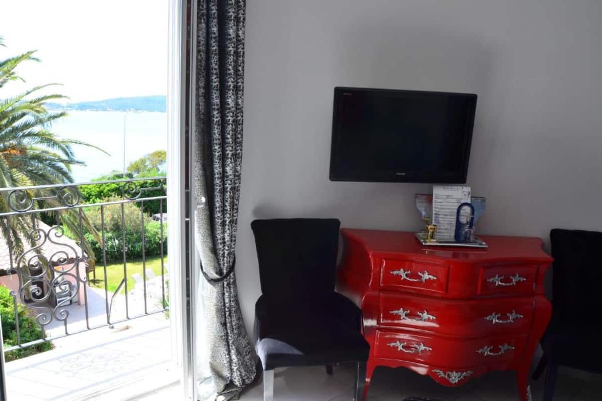 Una habitación con televisión en la pared, un aparador rojo y vista al mar desde un balcón.