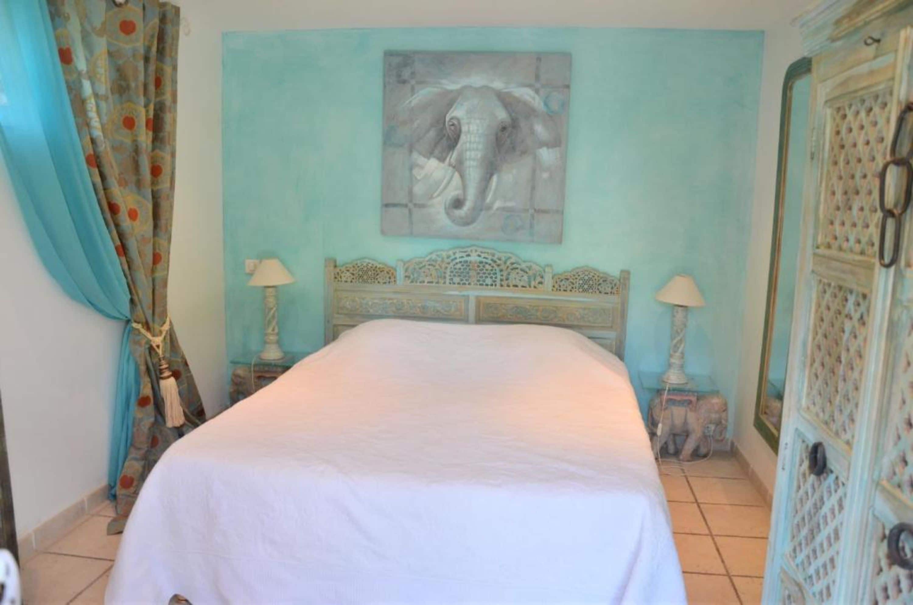 A bedroom with a bed, two bedside lamps, a wall painting of an elephant, and a patterned curtain.