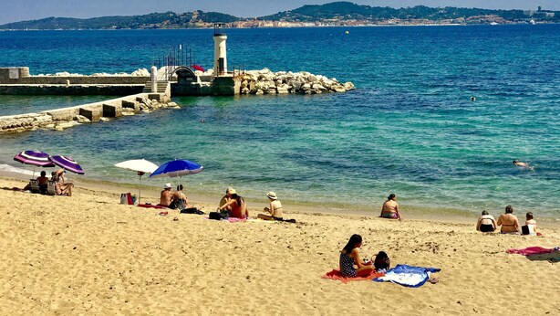 A beach with people sunbathing and swimming, a lighthouse, and a rocky breakwater. - Hôtel Villa des Anges (Grimaud)