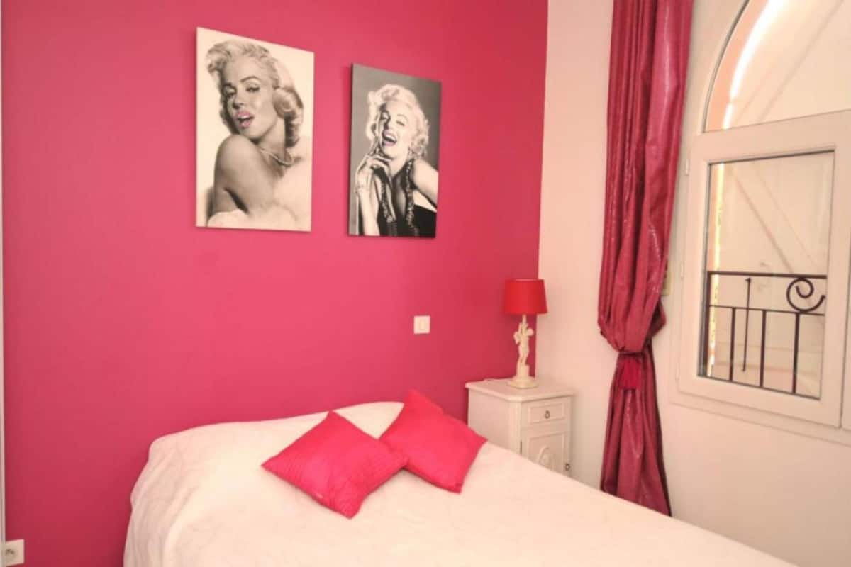 Un dormitorio con cama, dos carteles en la pared, una mesita de noche con lámpara y una ventana con cortinas.