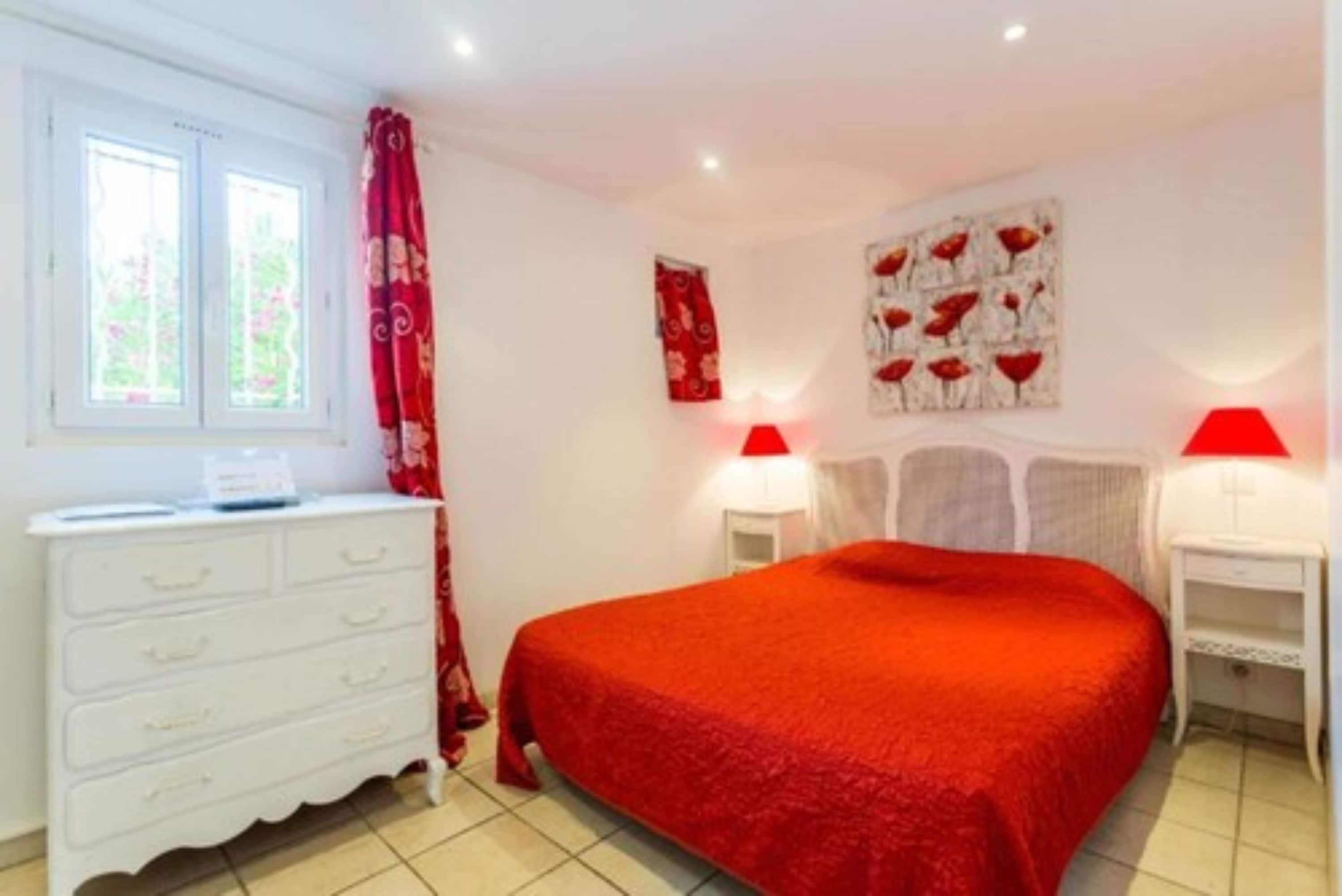 A bedroom with a red bedspread, white dresser, two bedside lamps, and a window with curtains.