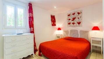 A bedroom with a red bedspread, white dresser, two bedside lamps, and a window with curtains.