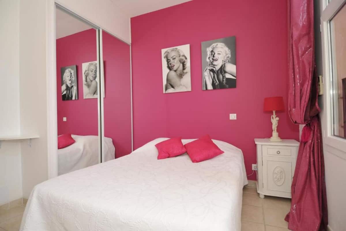 Un dormitorio con pared rosa, una cama con ropa de cama blanca y una mesita de noche con lámpara.