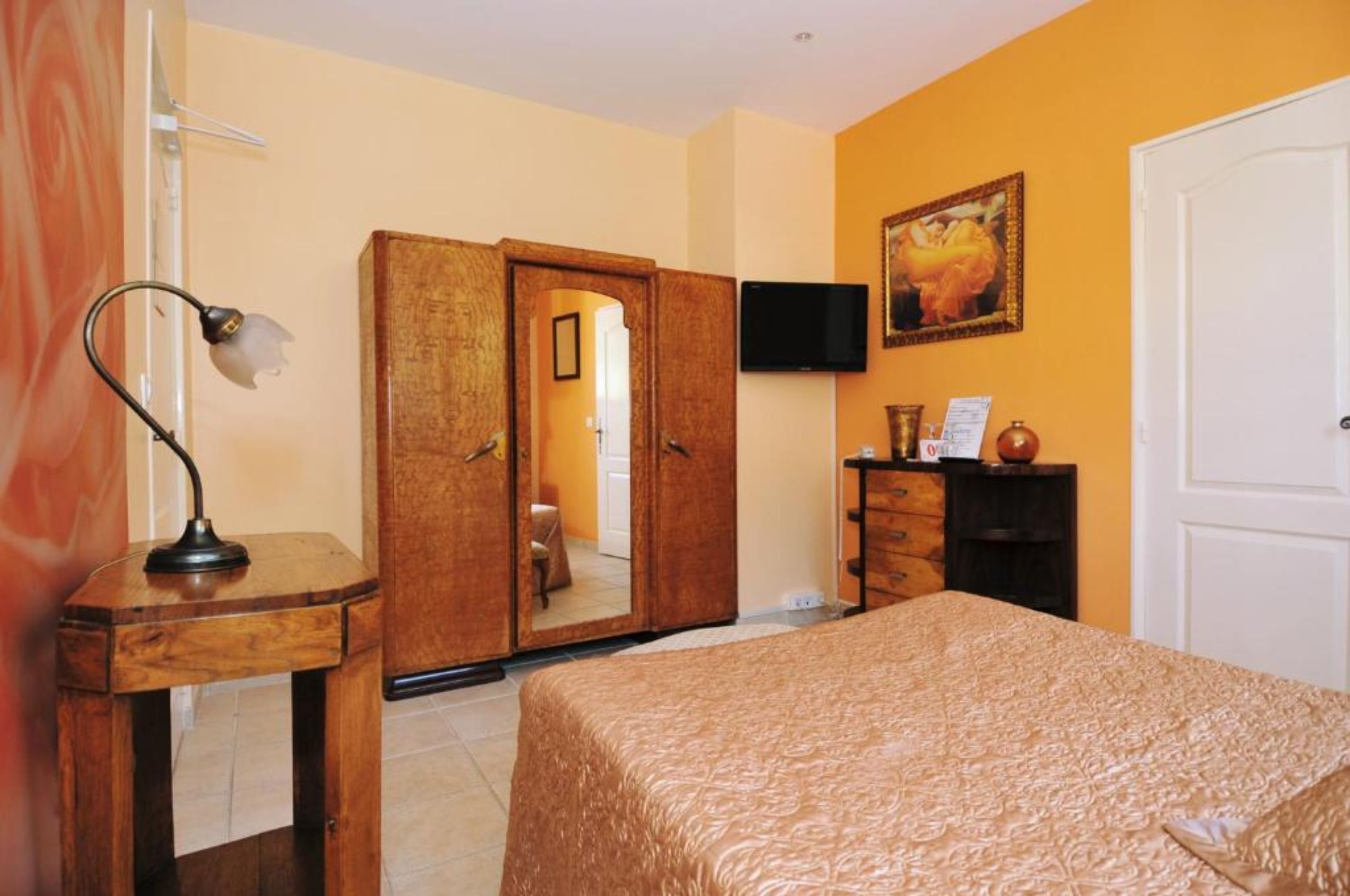 Une chambre d’hôtel avec un lit, une armoire en bois, un bureau avec une lampe, un téléviseur fixé au mur et un tableau.