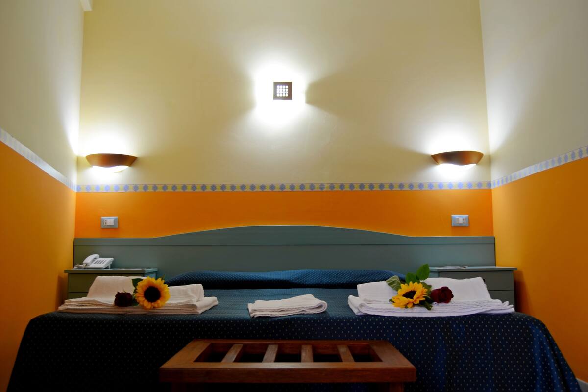 A hotel room with a bed, two bedside tables, a nightstand, a wooden bench, and a wall-mounted light fixture.