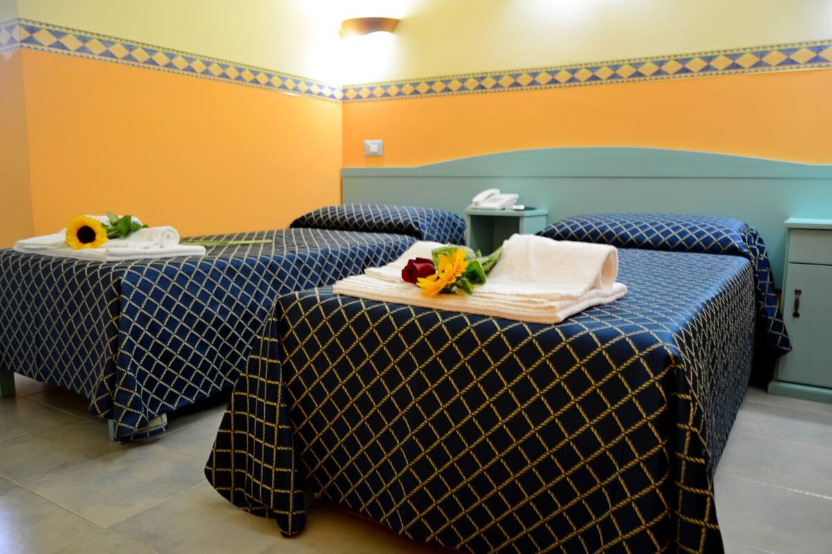 A hotel room with two beds, each with a blue and gold patterned bedspread, a nightstand with a telephone, and a small trash bin.