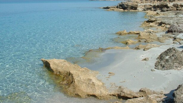 Un paisaje costero con agua azul cristalina, costa rocosa y colinas a lo lejos.