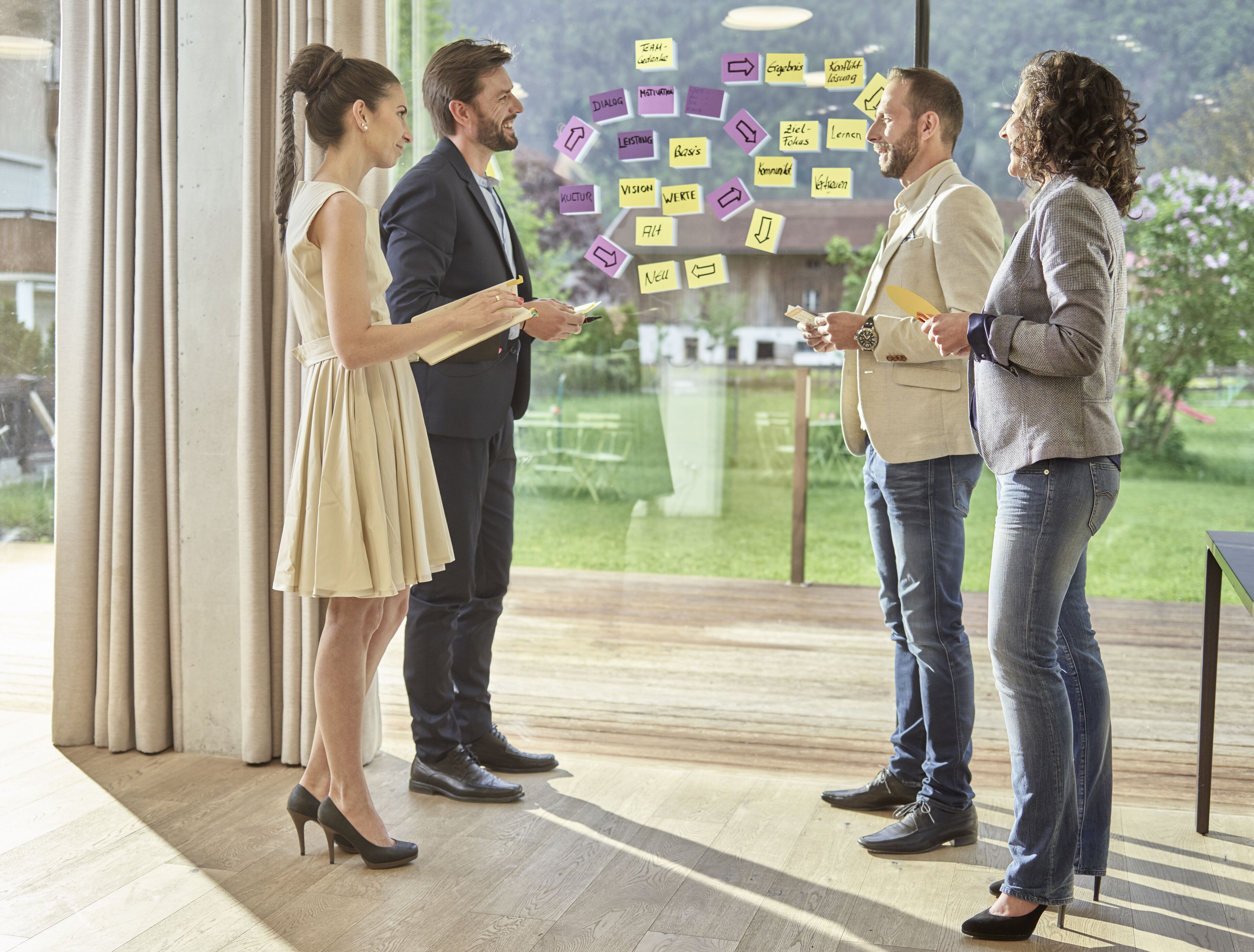 Cuatro personas en una oficina moderna con ventanas amplias y un muro de cristal, conversando sobre ideas escritas en notas adhesivas.