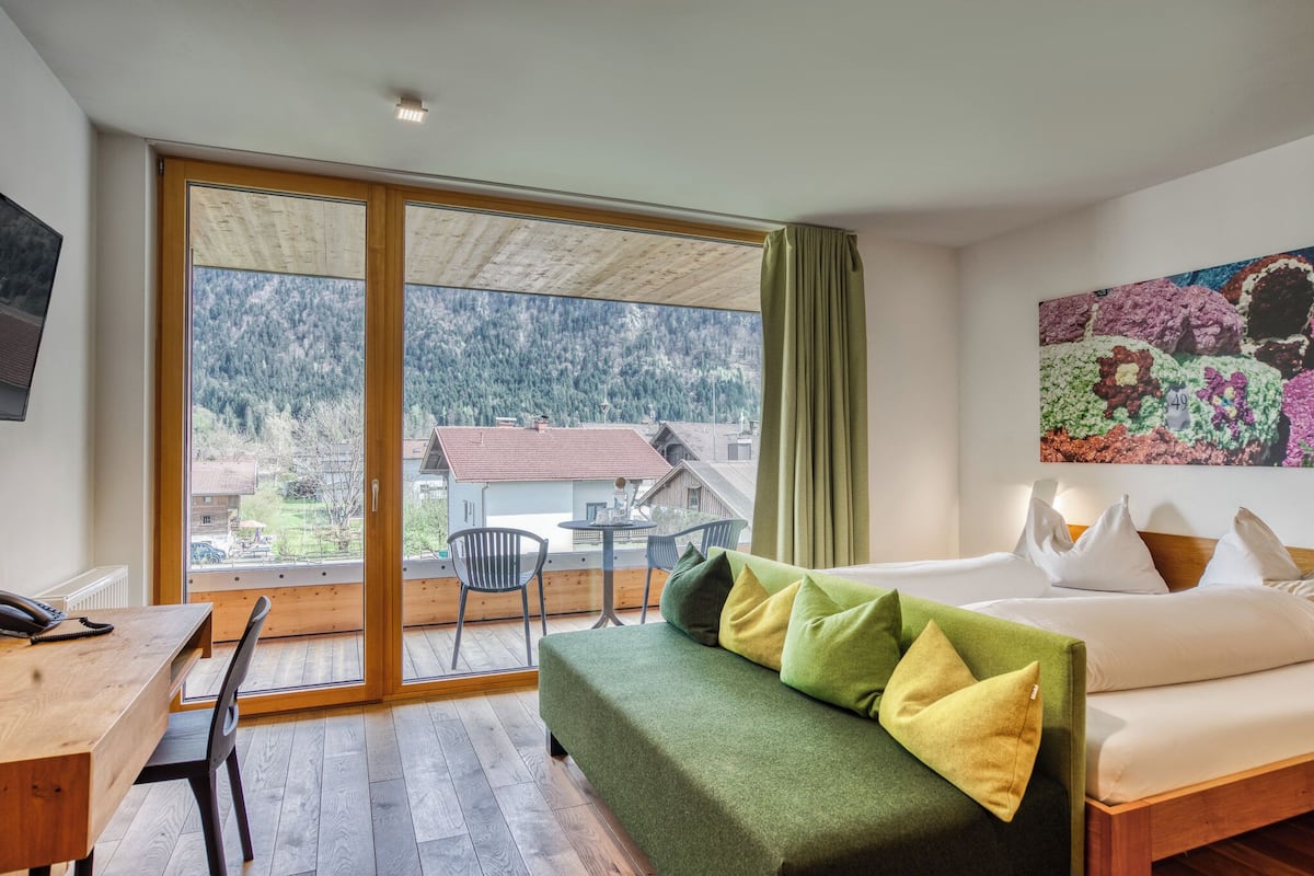 A room with a bed, a desk, and a chair. There is a window with a view of houses and trees.