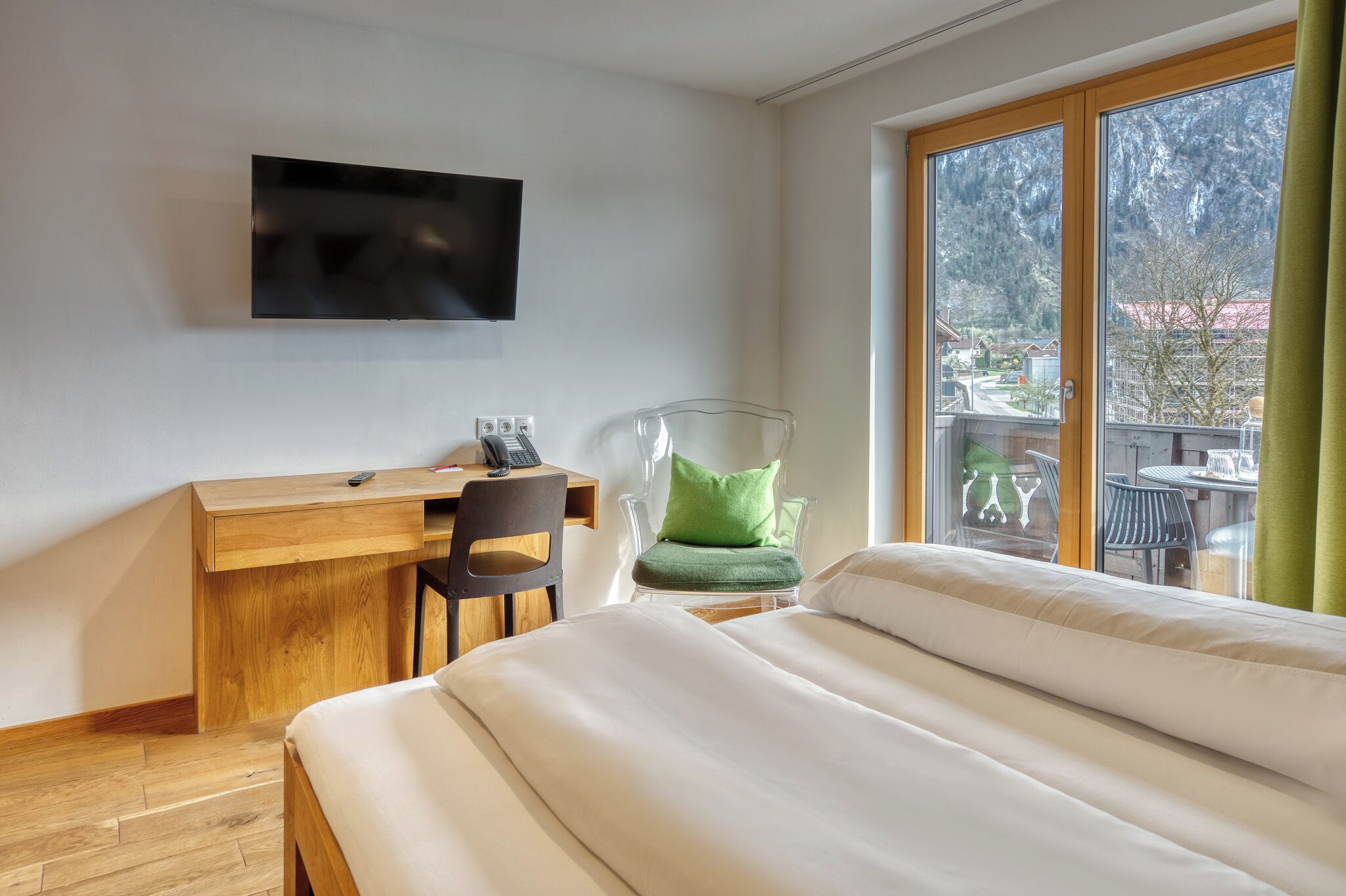 Habitación de hotel con cama, escritorio con silla, televisor montado en la pared y un ventanal con vistas a una montaña y un pueblo.