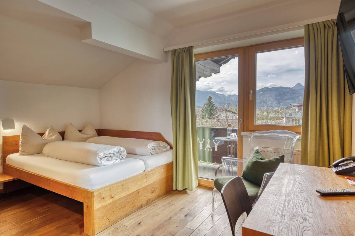 A room with a wooden bed, a dining table, and a view of mountains through large windows.