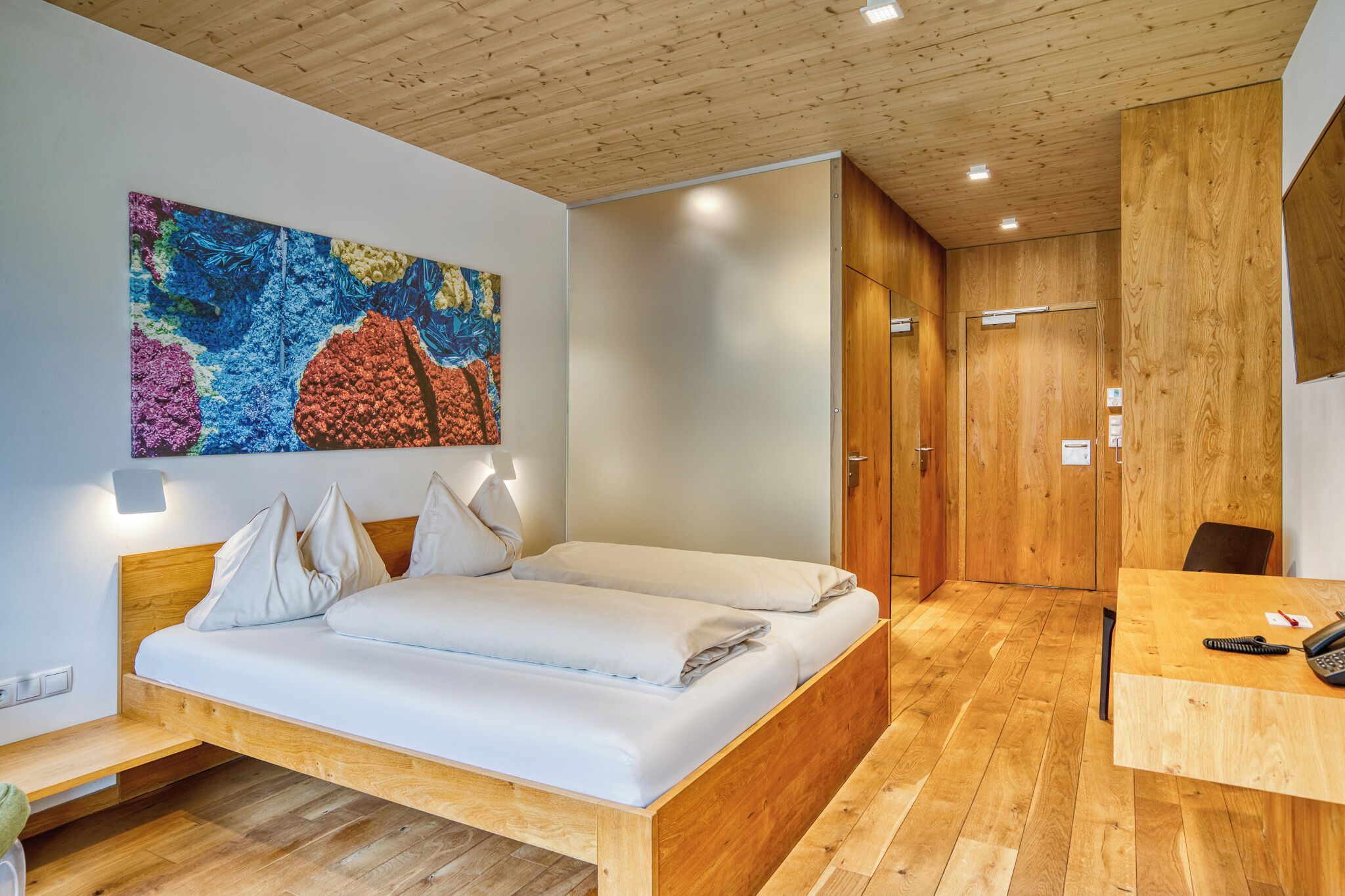 A modern hotel room with a wooden bed, a desk, and a large abstract painting on the wall.