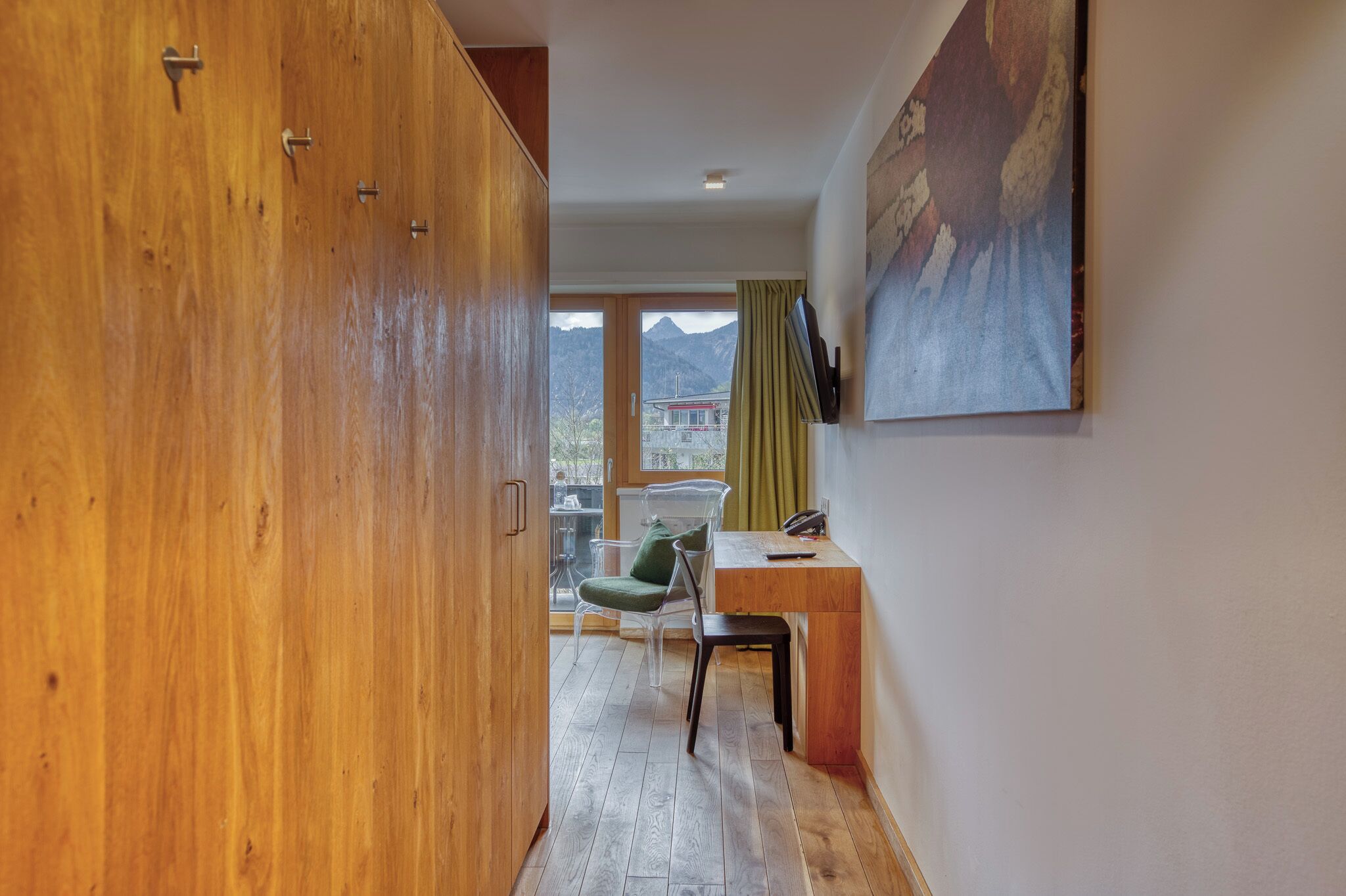 A hallway with wooden flooring, a desk with a chair, a painting on the wall, and a view of mountains through large windows.