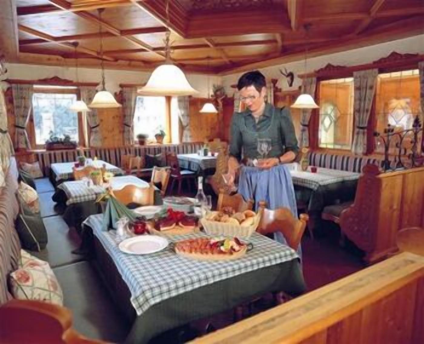 Interior de madeira com mesas preparadas para refeições, uma pessoa ao lado da mesa e um espaço de estar com bancos.