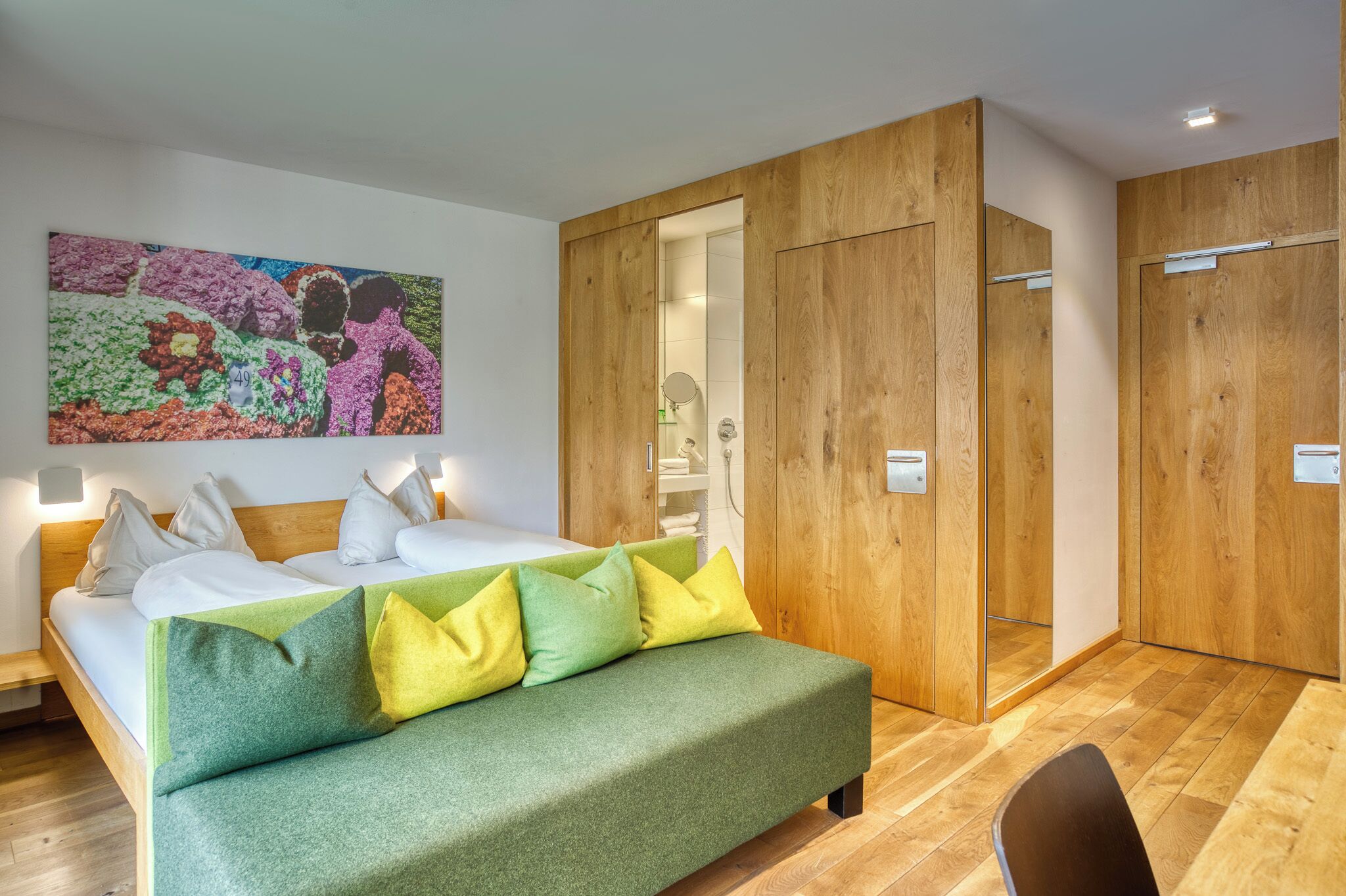 A modern hotel room with a bed, a green sofa, wooden wardrobe, and a colorful abstract painting on the wall.