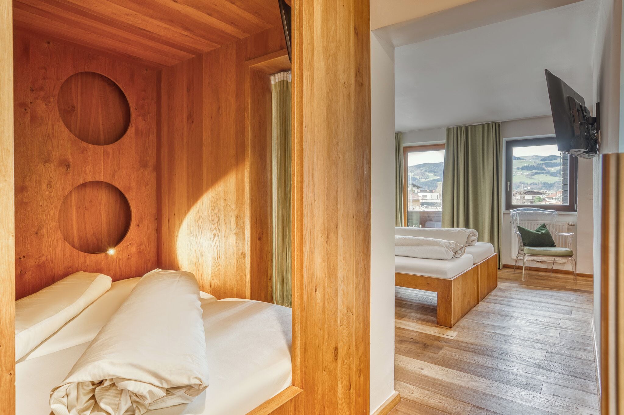 Een houten kamer met een bed en een slaapbank, een aan de muur bevestigde televisie, en uitzicht op buiten.
