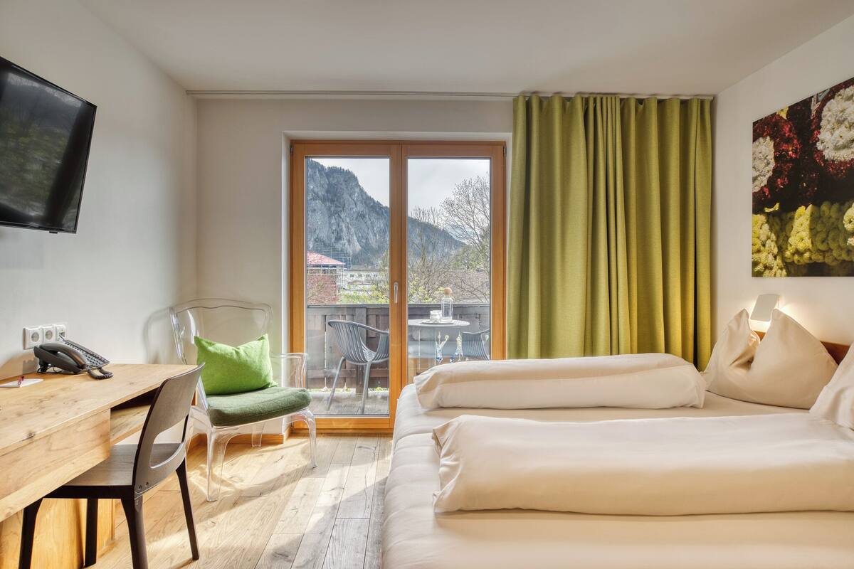 A hotel room with a wooden desk, a chair, a bed with white linens, a television, a sliding glass door to a balcony, and a green curtain.