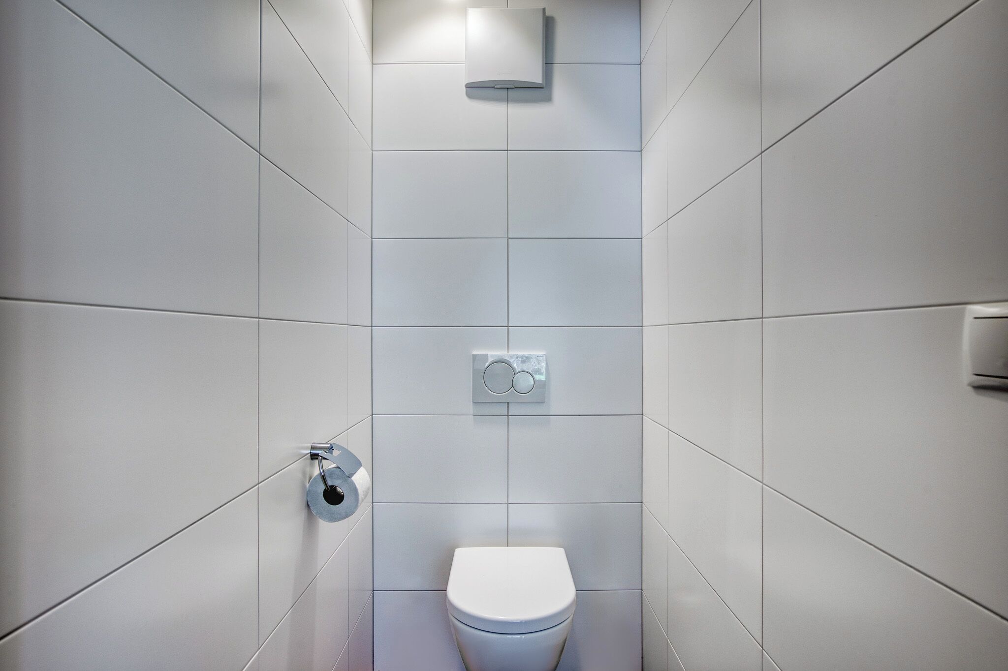 Un baño moderno con azulejos blancos, un inodoro, un dispensador de papel higiénico en la pared y un perchero para toallas.