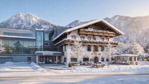 Un hotel con fachada alpina tradicional, un edificio moderno de vidrio contiguo y un paisaje montañoso nevado de fondo.