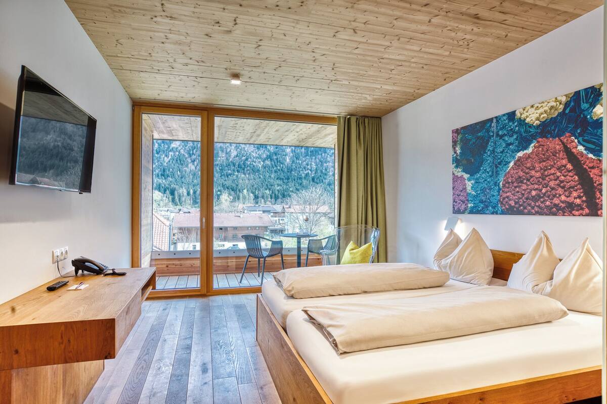 A modern hotel room with a wooden ceiling, a large window with a view of a forest, a bed with white linens, a desk with a phone, and a painting on the wall.