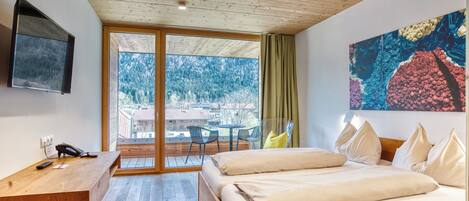Una habitación de hotel moderna con techo de madera, un ventanal con vistas a un bosque, cama con ropa blanca, escritorio con teléfono y un cuadro en la pared.