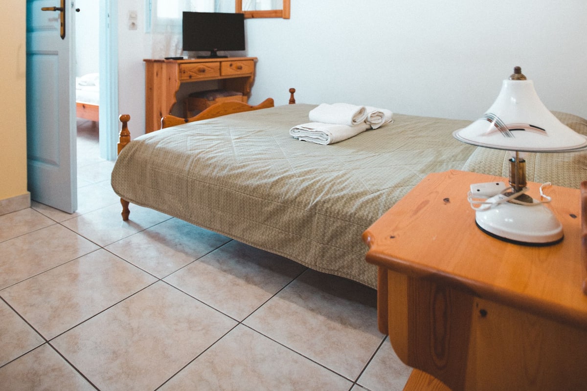 A bedroom with a bed, a wooden nightstand, a desk, a lamp, a mirror, a television, and a door leading to another room.