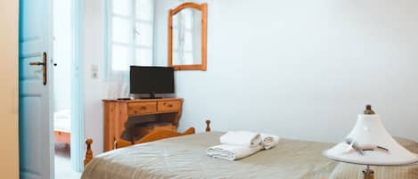 Un dormitorio con una cama, una mesita de noche de madera, un escritorio, una lámpara, un espejo, un televisor y una puerta que da a otra habitación.