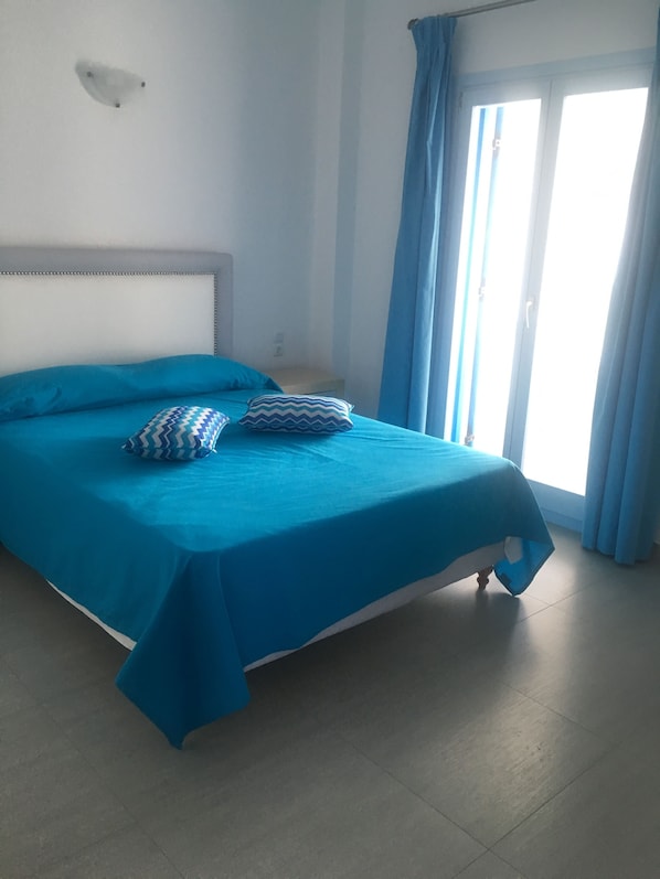 A room with a bed, a headboard, a window with curtains, and a wall-mounted light fixture.
