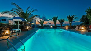 Piscina com espreguiçadeiras e guarda-sóis, rodeada por palmeiras e vista para o pôr do sol.
