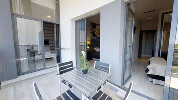 A modern outdoor balcony with a table and chairs, a potted plant, and a sliding glass door leading to an interior space.