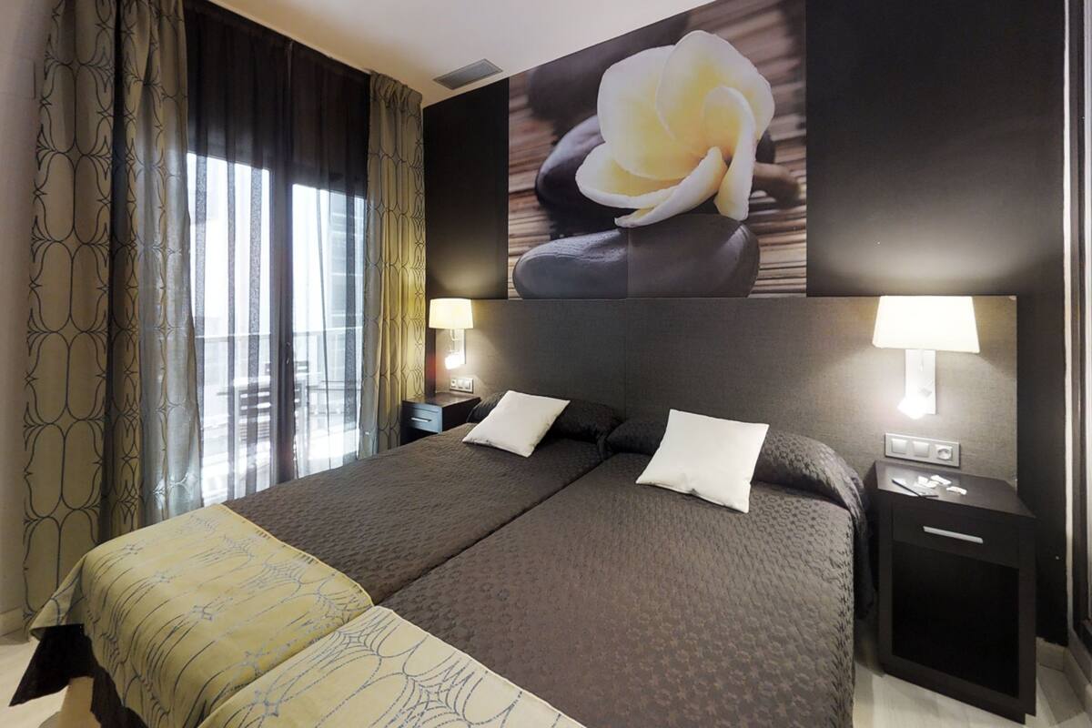 A double bed with two pillows, a bedside table, a nightstand, a wall-mounted lamp, and a large window with curtains.