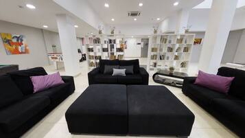 A spacious living area with black sofas and ottomans, a central coffee table, and a bookshelf filled with books.