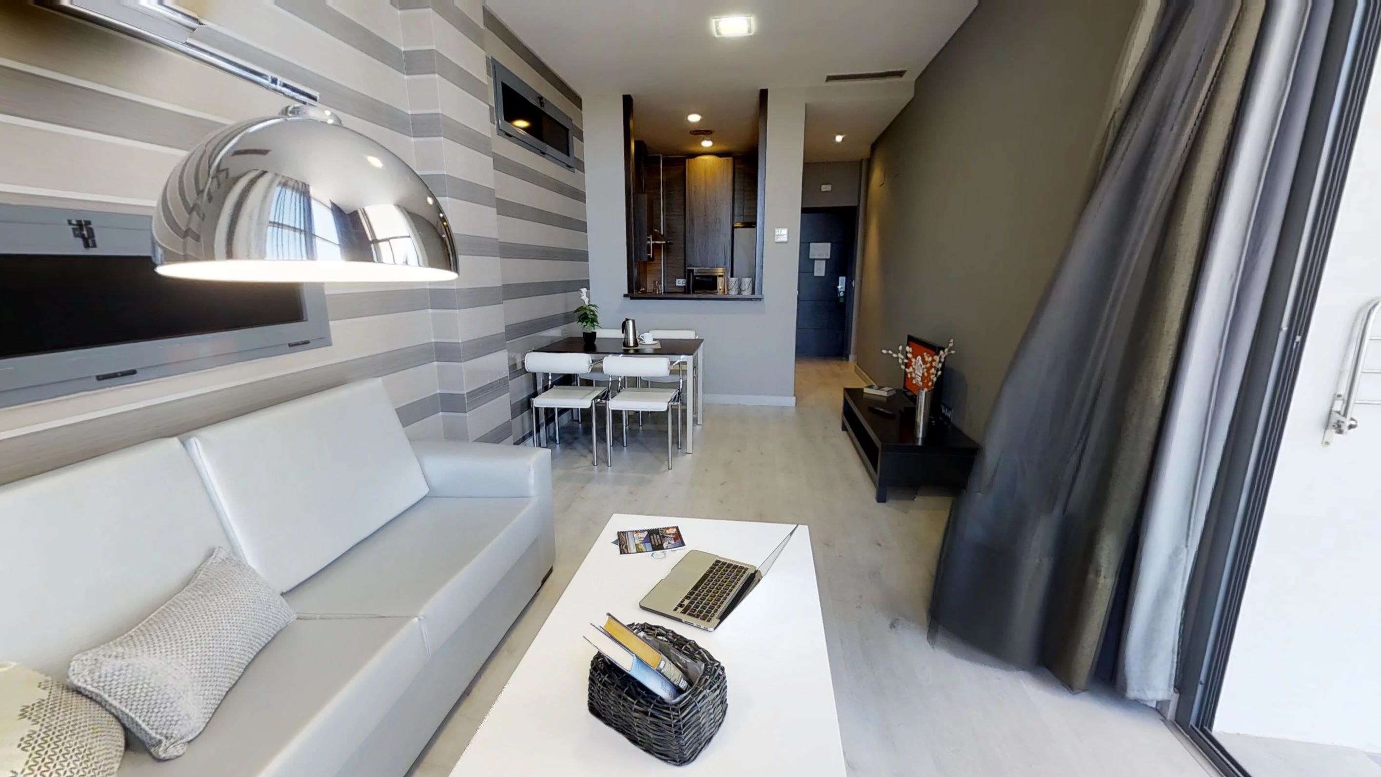 A modern living room with a white sofa, a coffee table with magazines, and a dining area with a table and chairs.