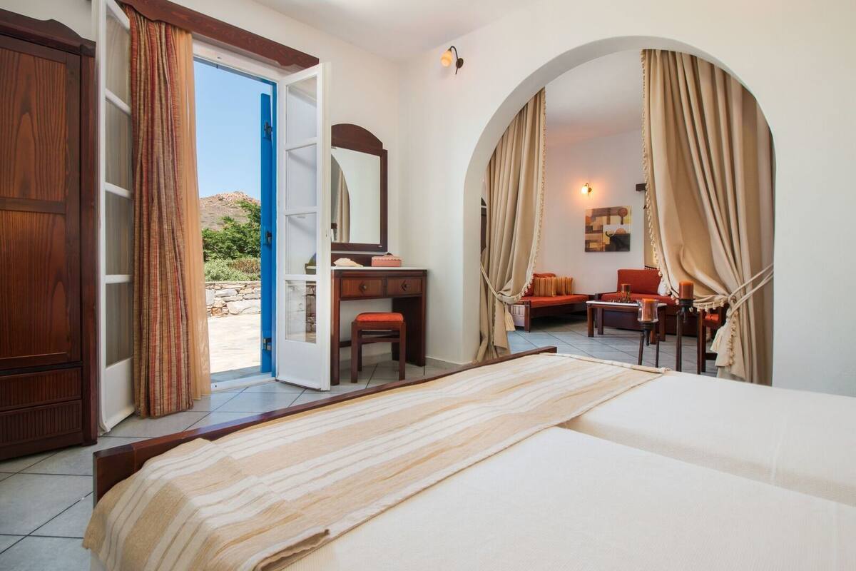 A hotel room with a bed, a desk, a chair, and a sofa. There is a window with curtains, a mirror, and a wall-mounted light.